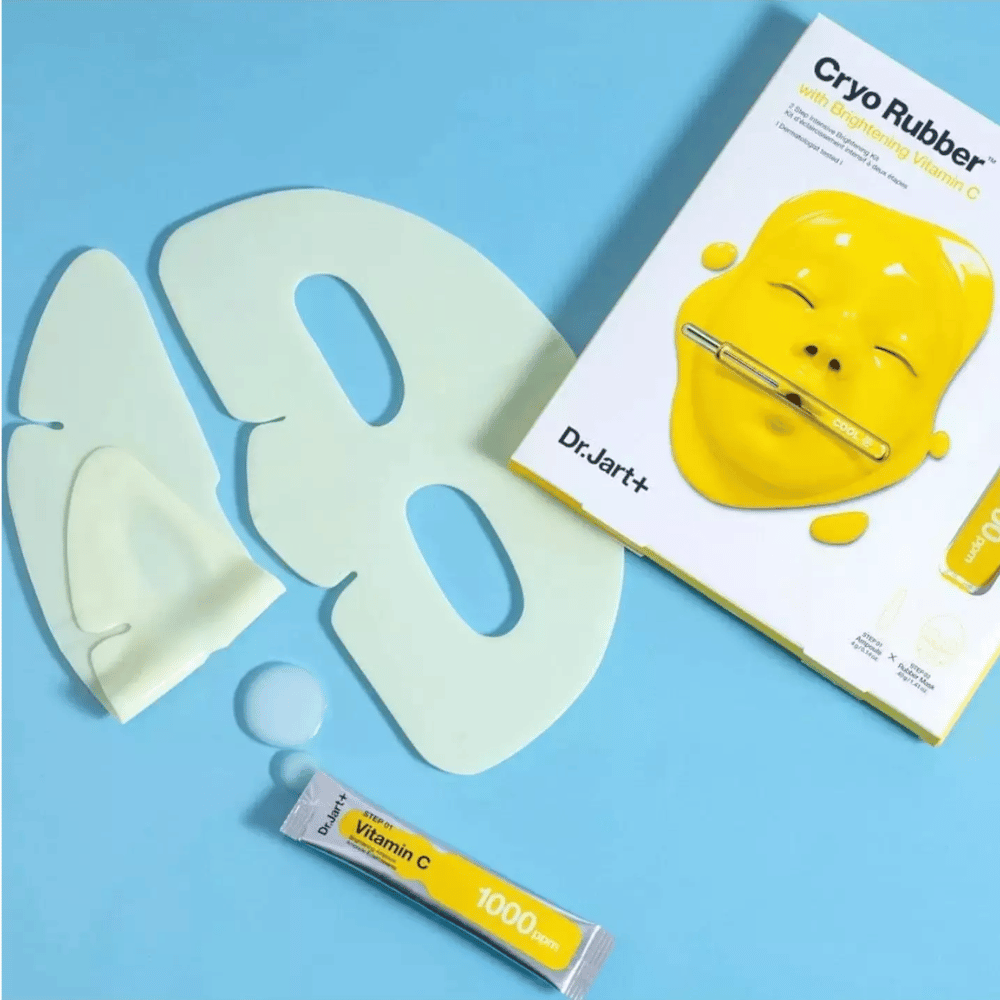 Obraz Dr.Jart+'s 'Cryo Rubber™ z Rozjaśniającą Witaminą C' produkt kosmetyczny. Na niebieskim tle widać białą gumową maskę na twarz, małą tubkę oznaczoną 'Witamina C 1000ppm' oraz pudełko z nazwą produktu i obrazem żółtej maski.