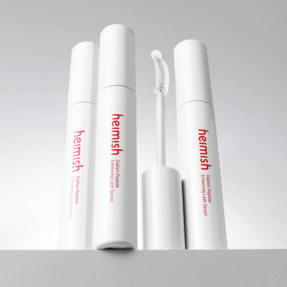 Kilka opakowań Heimish Dailism Peptide Enhancing Lash Serum jest pokazanych razem z wygiętą szczoteczką aplikatora. Obraz podkreśla elegancki design produktu oraz precyzyjną szczoteczkę ułatwiającą aplikację.