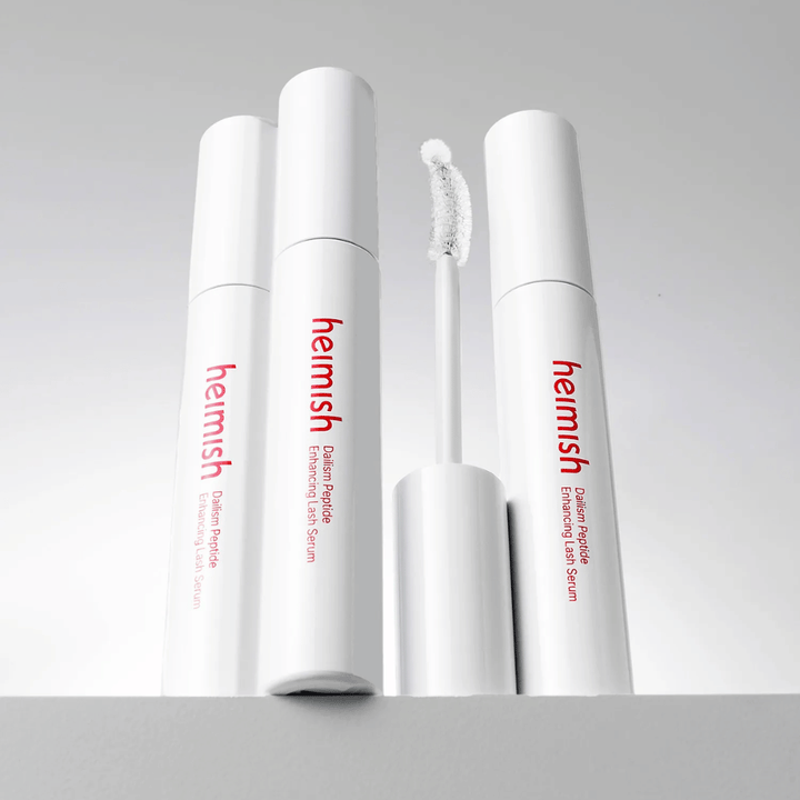 Kilka opakowań Heimish Dailism Peptide Enhancing Lash Serum jest pokazanych razem z wygiętą szczoteczką aplikatora. Obraz podkreśla elegancki design produktu oraz precyzyjną szczoteczkę ułatwiającą aplikację.