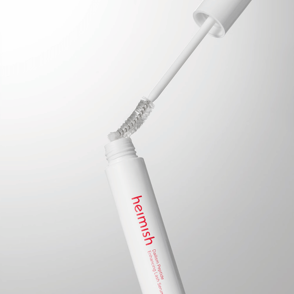 Zbliżenie na aplikator Heimish Dailism Peptide Enhancing Lash Serum. Szczoteczka jest wyciągana z białego pojemnika i pokazuje przezroczystą formułę serum, którą nakłada się na rzęsy.