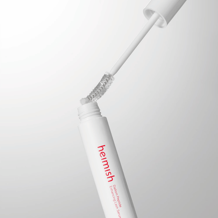Zbliżenie na aplikator Heimish Dailism Peptide Enhancing Lash Serum. Szczoteczka jest wyciągana z białego pojemnika i pokazuje przezroczystą formułę serum, którą nakłada się na rzęsy.