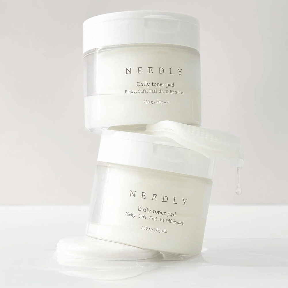 Dwa pojemniki z Needly Daily Toner Pad ułożone jeden na drugim. Pojemniki są białe z minimalistycznym tekstem i zawierają po 60 toników każdy. Jeden tonik częściowo wystaje z górnego pojemnika, a płyn kapie.