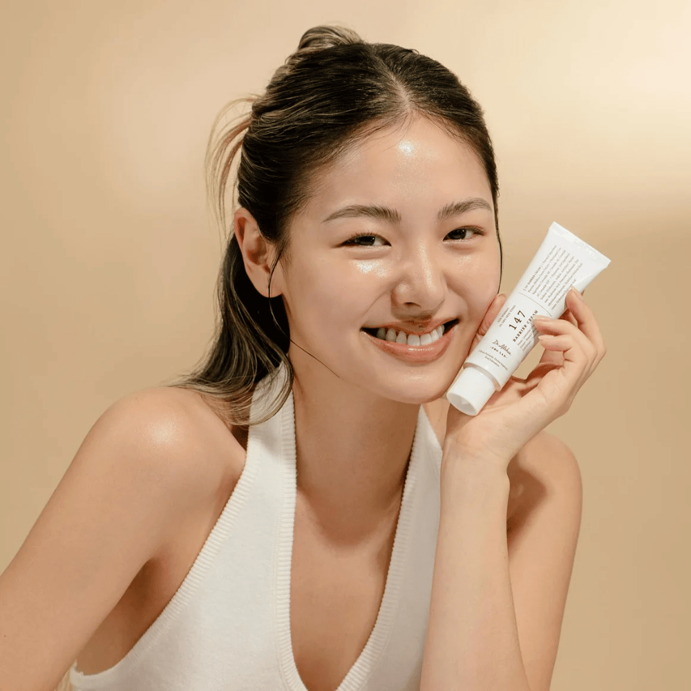 Modelka z promienną skórą uśmiecha się i trzyma Dr. Althea 147 Barrier Cream – koreański krem do twarzy do bariery skórnej i nawilżania.