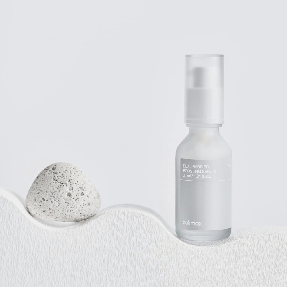 Celimax Dual Barrier Boosting Serum umieszczony na tkaninie z kamieniem, spokojny obraz produktu sygnalizujący nawilżenie i wzmocnioną barierę skórną.