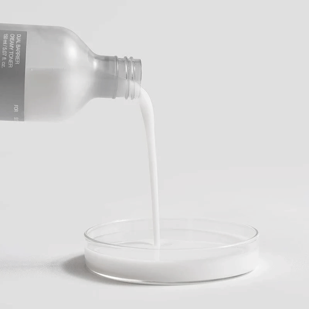 Celimax Dual Barrier Creamy Toner hälls försiktigt från flaskan ner i en rund laboratoriebrunn, med sin mjölkiga och näringsrika formula tydligt i fokus.
