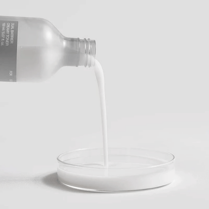 Celimax Dual Barrier Creamy Toner hälls försiktigt från flaskan ner i en rund laboratoriebrunn, med sin mjölkiga och näringsrika formula tydligt i fokus.