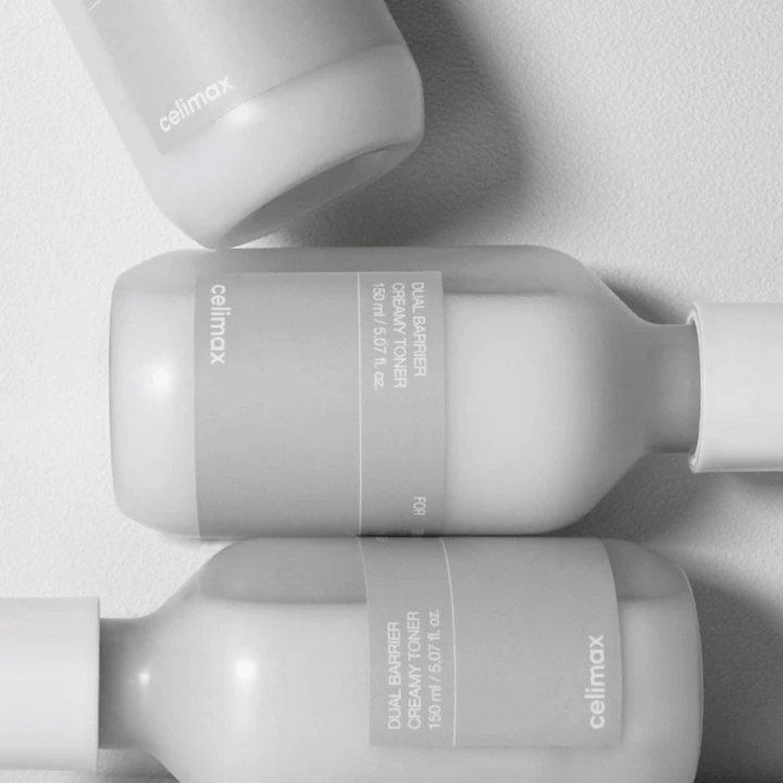 Tre liggande flaskor av Celimax Dual Barrier Creamy Toner i en minimalistisk design med grå etikett och mjukt ljus – perfekt för känslig och torr hud.