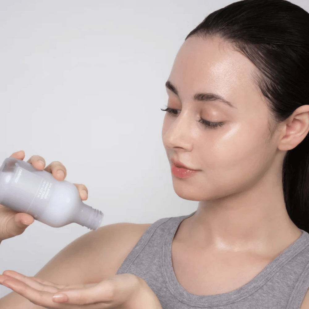 Kobieta z błyszczącą, nawilżoną skórą nakłada Celimax Dual Barrier Creamy Toner poprzez wylewanie produktu na dłoń przed swoją rutyną pielęgnacyjną.