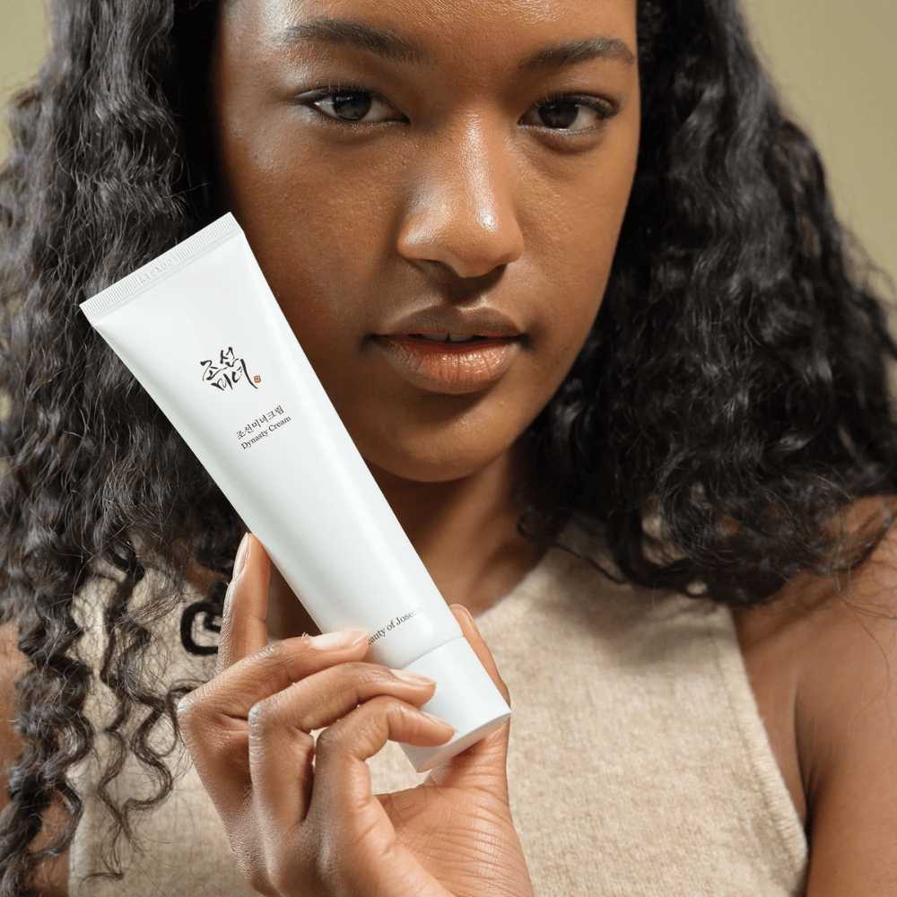 Kobieta z kręconymi włosami trzyma tubkę "Beauty of Joseon Dynasty Cream" blisko swojej twarzy, co podkreśla produkt. Krem, prezentowany w eleganckiej białej tubce, jest zaprojektowany, aby odżywiać i odmładzać skórę, co podkreśla jego atrakcyjność dla różnych typów skóry.