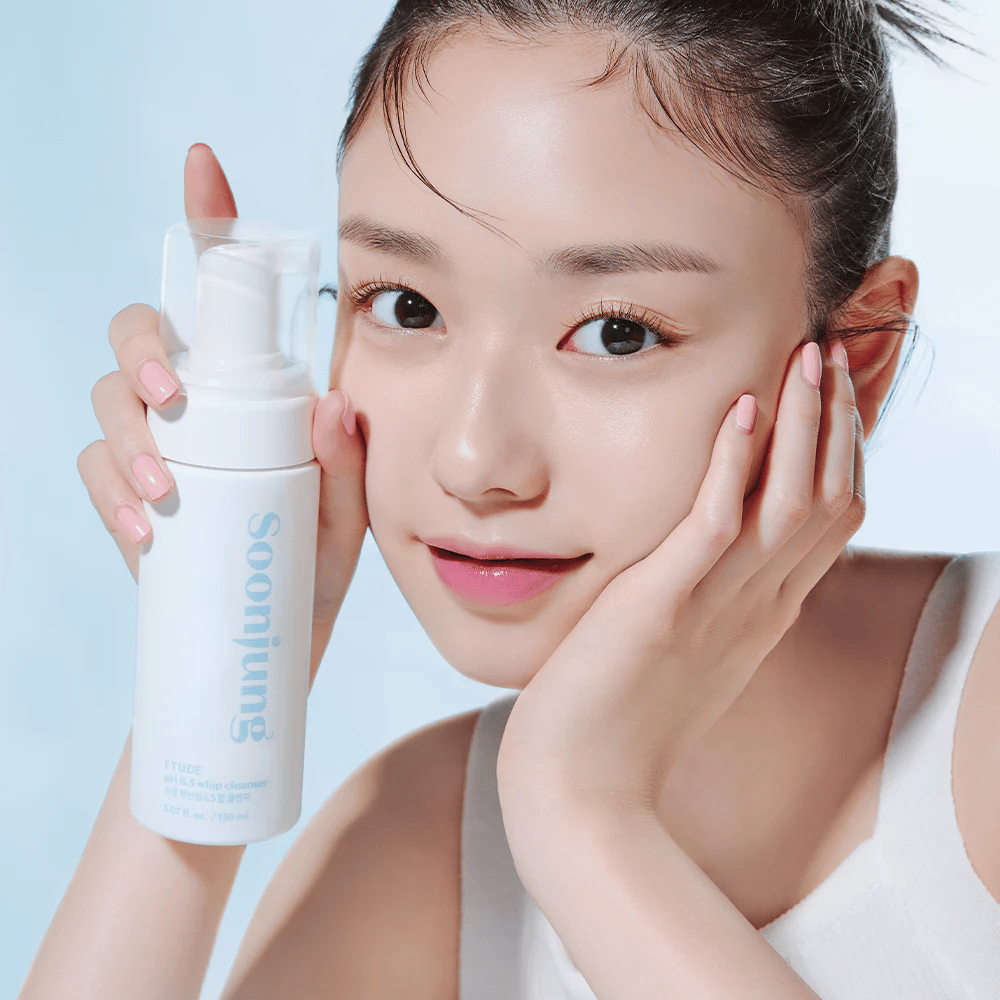 Młoda kobieta z jasną cerą trzyma białą butelkę z pompką z napisem "Soon Jung pH 6.5 Whip Cleanser" od ETUDE. Uśmiecha się do kamery z lekkim makijażem i świeżym wyglądem na jasnoniebieskim tle.
