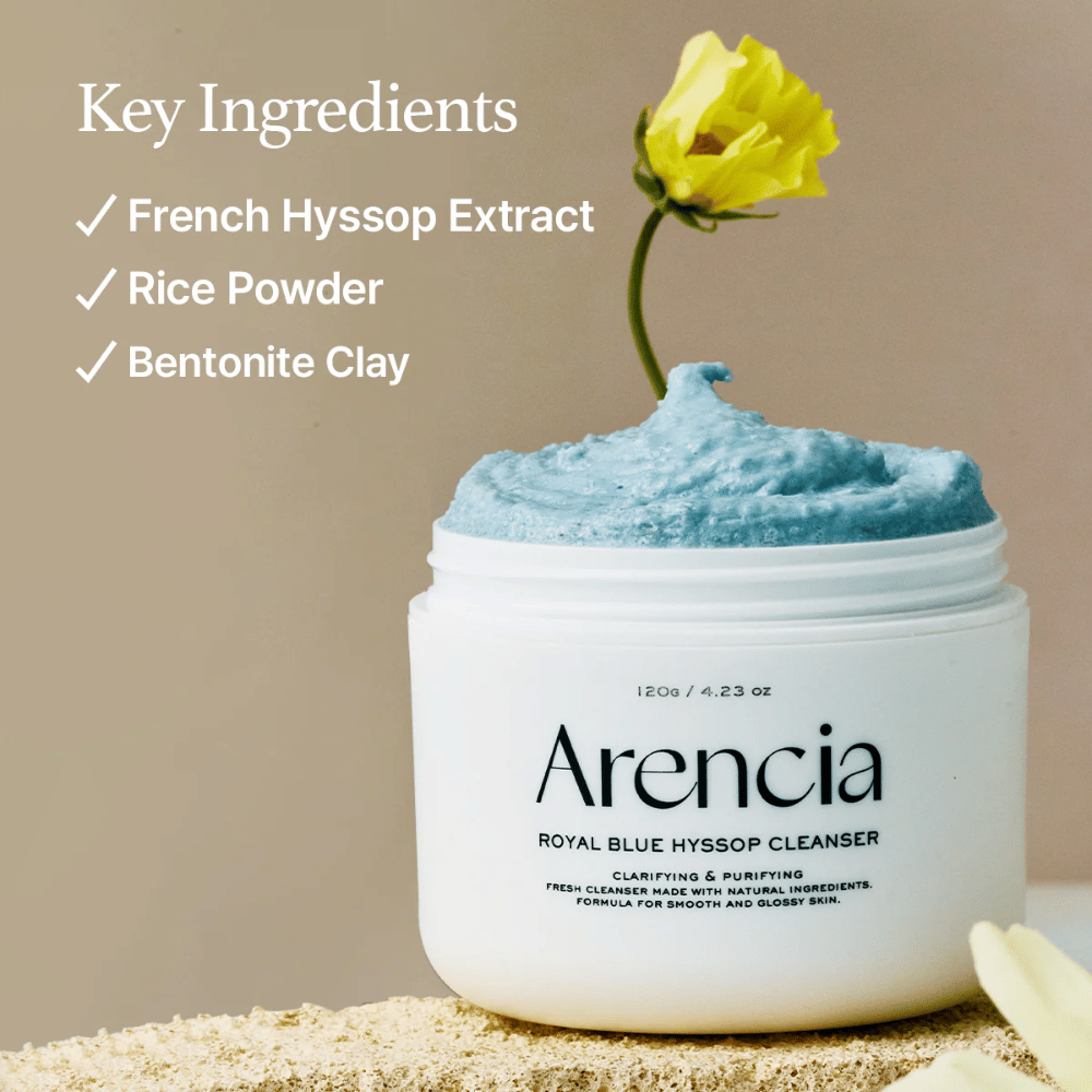 Zdjęcie słoika "Arencia Royal Blue Hyssop Cleanser" wypełnionego niebieskim kremem czyszczącym. Żółty kwiat wystaje z kremu. Tekst na zdjęciu podkreśla trzy kluczowe składniki: ekstrakt z francuskiego hyzopu, puder ryżowy i glinkę bentonitową.