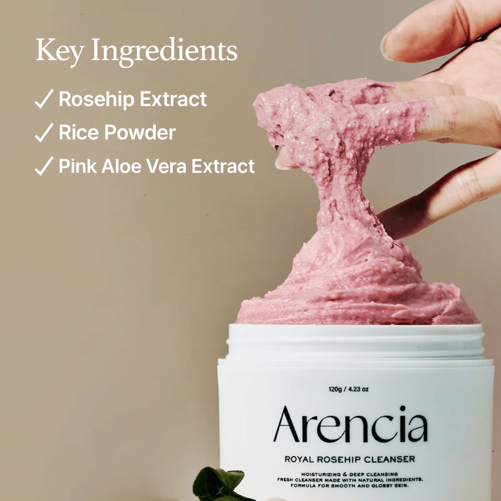 Ręka podnosi różową, puszystą konsystencję oczyszczającą ze słoika Arencia Royal Rosehip Cleanser, z listą składników po lewej: ekstrakt z dzikiej róży, proszek ryżowy i różowy ekstrakt z aloesu.