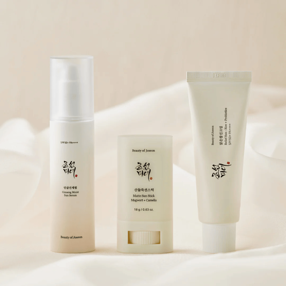 Trzy produkty do ochrony przeciwsłonecznej od Beauty of Joseon – Ginseng Moist Sun Serum, Matte Sun Stick i Relief Sun Rice + Probiotics – stoją w rzędzie na kremowym materiale w miękkim świetle.