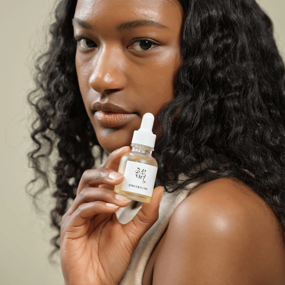 Kobieta z kręconymi włosami trzyma butelkę Beauty of Joseon Glow Serum z pipetą, nawilżający i rozświetlający produkt do pielęgnacji skóry.