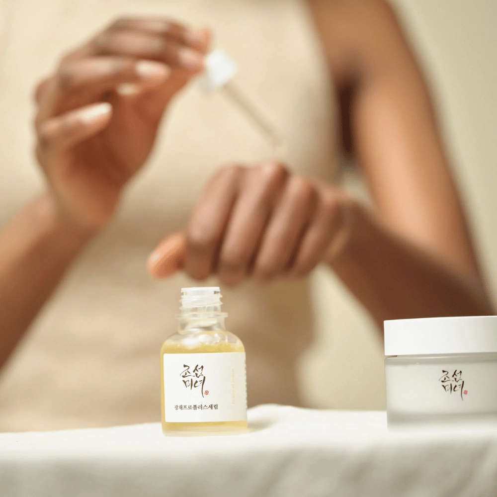 Osoba aplikująca Beauty of Joseon Glow Serum z pipetą z szklanej butelki, z słoikiem z tej samej serii obok na stole.