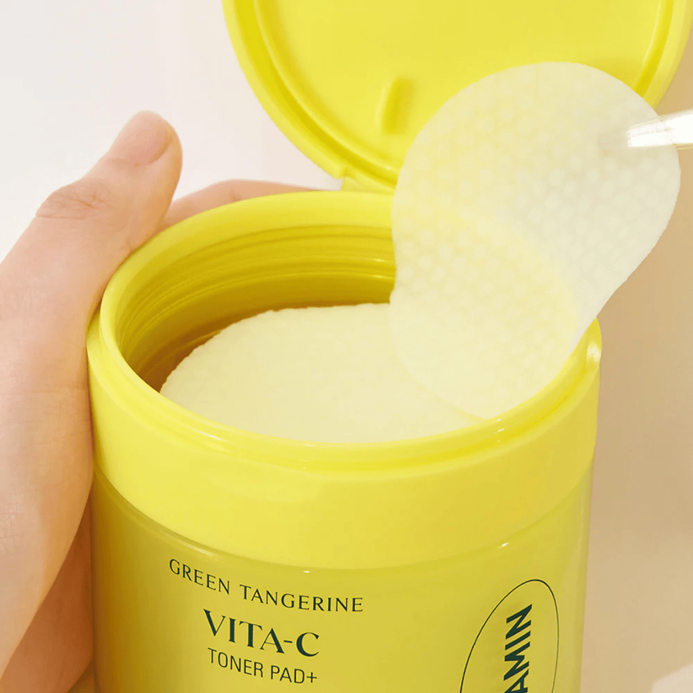 Ręka używa plastikowej pęsety, aby podnieść teksturowaną płytkę tonizującą z jasnożółtego pojemnika oznaczonego "GREEN TANGERINE VITA-C TONER PAD+" marki "gooda".