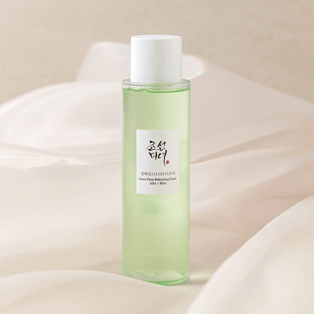 Butelka z "Green Plum Refreshing Toner" AHA + BHA stoi na miękkim, jasnym tle. Produkt ma zielony płyn, który odzwierciedla świeżość i naturalność. Etykieta jest biała z czarnymi i czerwonymi elementami, co daje elegancki i czysty wygląd.