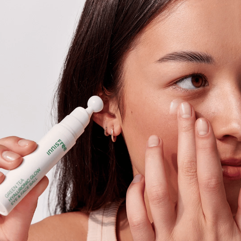 Osoba nakłada Innisfree Green Tea Hyaluronic Glow Eye & Face Ball na swoją skórę pod oczami. Produkt ma białą, okrągłą część aplikacyjną i znajduje się w białej butelce z zielonym napisem. Twarz użytkownika jest częściowo widoczna i skupia się na aplikacji produktu.