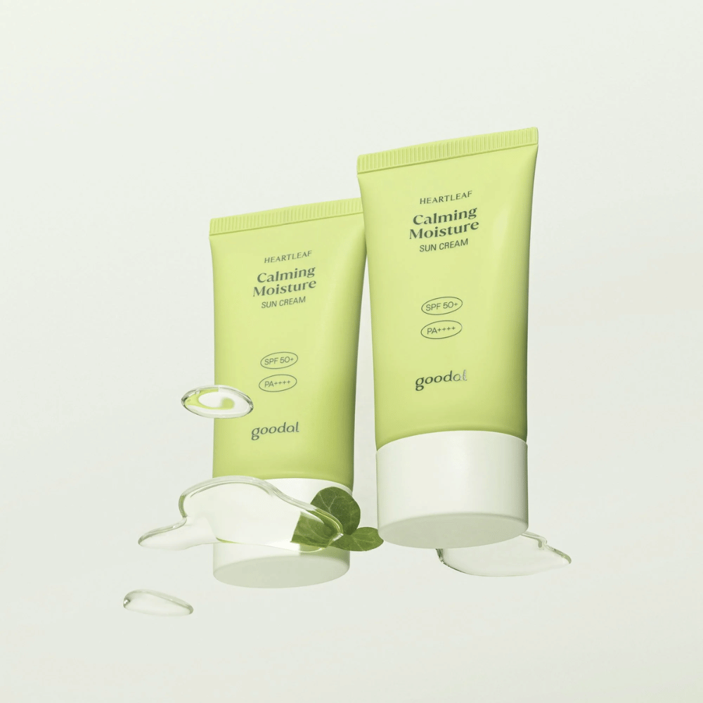 Dwie tubki Goodal Heartleaf Calming Moisture Sun Cream SPF50+ PA++++ otoczone liśćmi roślin i kroplami serum na jasnym tle.