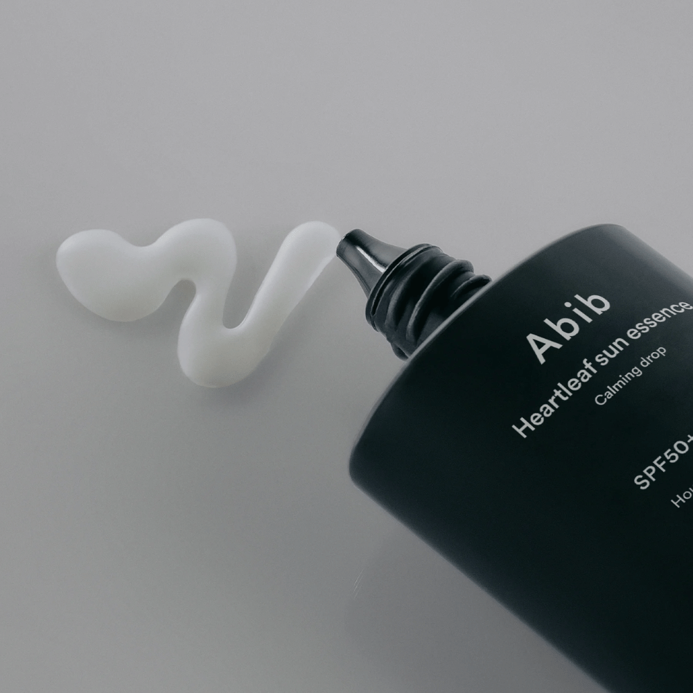 Zbliżenie na Abib Heartleaf Sun Essence Calming Drop z kremem przeciwsłonecznym wyciśniętym w białą, falistą linię na jasnej powierzchni.