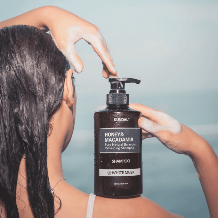 Kvinna med vått hår applicerar lödder och håller KUNDAL Honey & Macadamia Nature Shampoo White Musk mot havsbakgrund.