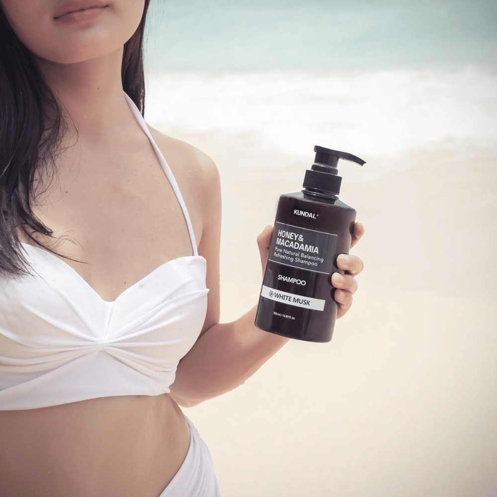 Kobieta w białym bikini trzyma KUNDAL Honey & Macadamia Nature Shampoo White Musk na plaży z morzem w tle.