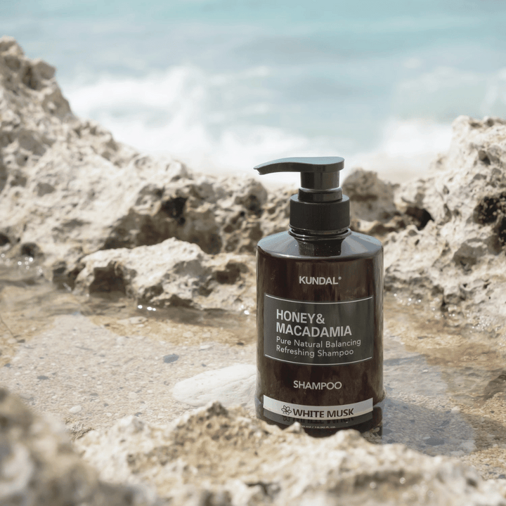 KUNDAL Honey & Macadamia Nature Shampoo White Musk placerad på klippor vid havet, en naturlig hårvårdsprodukt med fräsch doft.