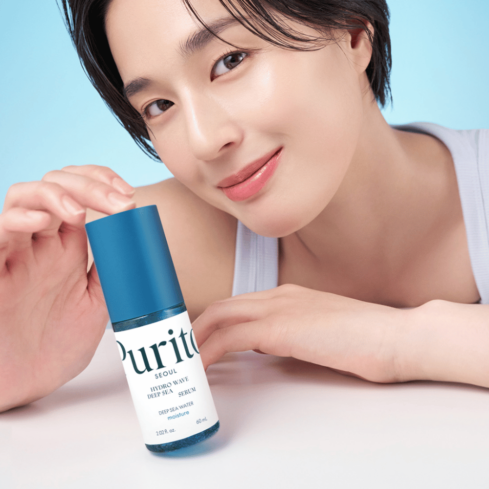 Kobieta z krótkimi włosami trzyma niebieskie serum od Purito, Hydro Wave Deep Sea Serum. Jej skóra wygląda promiennie i wilgotnie, co podkreśla produkt w centrum uwagi. Tło jest jasnoniebieskie, co daje świeże i czyste wrażenie. Produkt jest pogłębiony w pielęgnacji skóry.