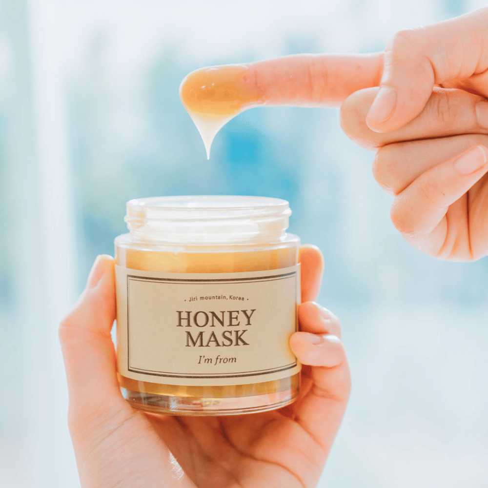 Ręka trzyma otwarty słoik "I'm from Honey Mask", a palec zanurza się w złotej masce na jasnym tle.