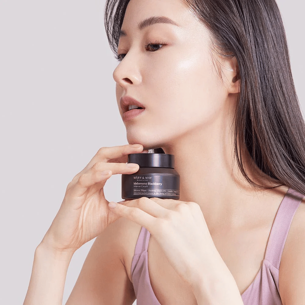 Kobieta trzyma słoik z MARY & MAY Idebenone Blackberry Intense Cream. Ma promienną cerę i lekko się uśmiecha, ubrana w bezrękawnik. Produkt to intensywny krem z idebenonem i kompleksem jeżynowym, przeznaczony do jędrności i nawilżenia skóry.
