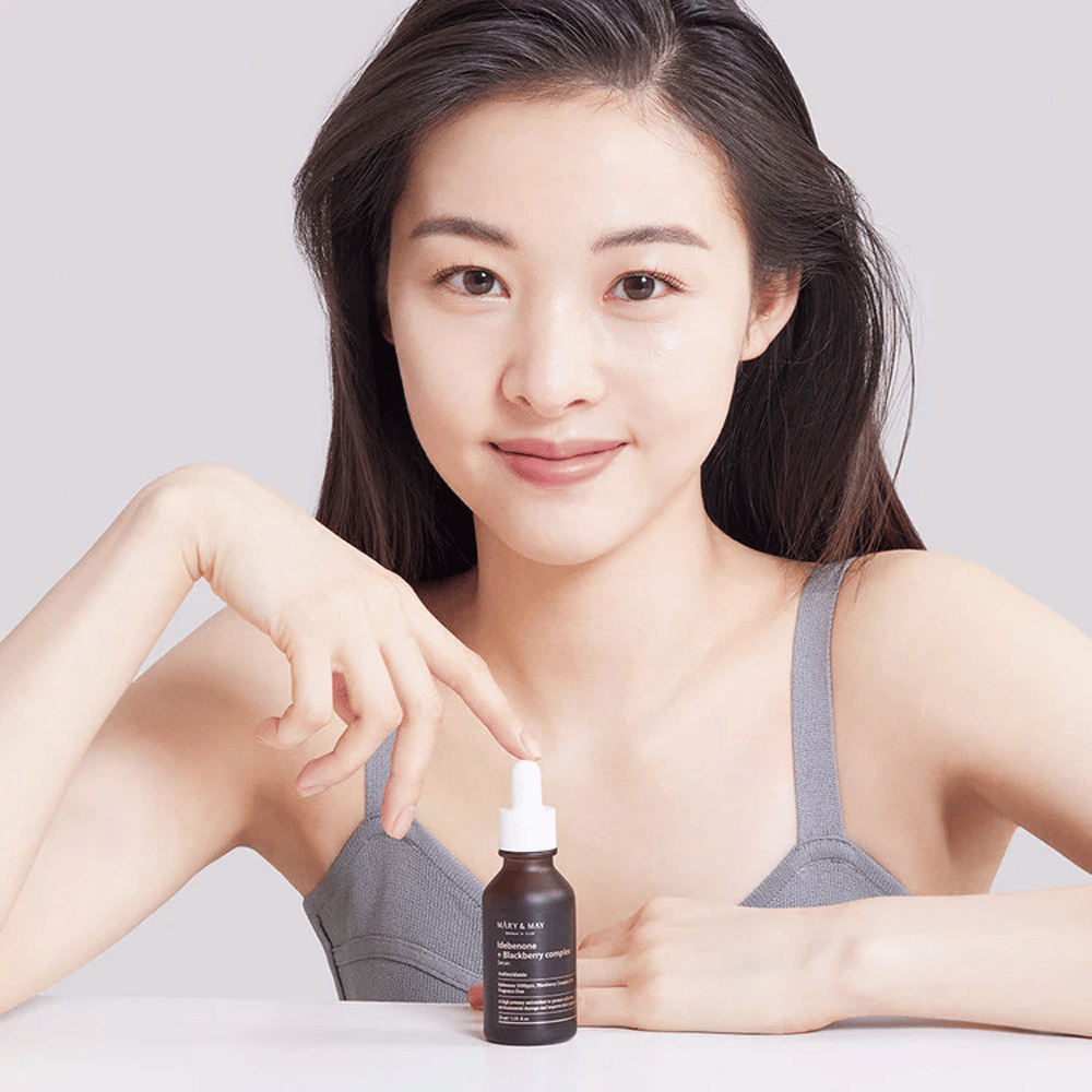 Kobieta z długimi, ciemnymi włosami trzyma w ręku MARY & MAY Idebenone + Blackberry Complex Serum, ciemną szklaną butelkę z pipetą. Ma radosny uśmiech i wskazuje na butelkę drugą ręką. Kobieta jest ubrana w szarą, bezrękawną bluzkę i wydaje się być w jasnym i prostym otoczeniu, co podkreśla skupienie produktu na pielęgnacji skóry i urodzie.