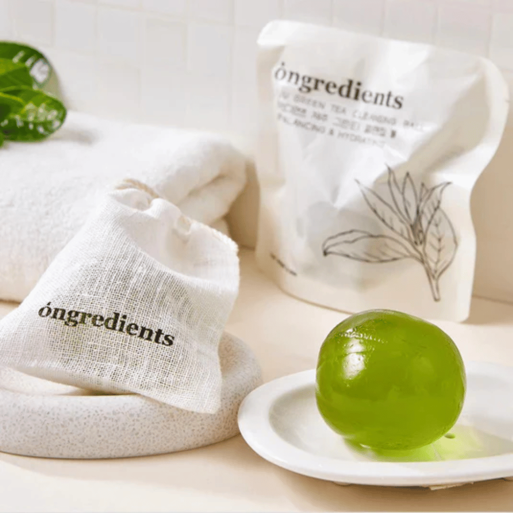 Óngredients Jeju Green Tea Cleansing Ball umieszczony na talerzu z opakowaniem i ręcznikiem – delikatne oczyszczanie z zieloną herbatą z Jeju.