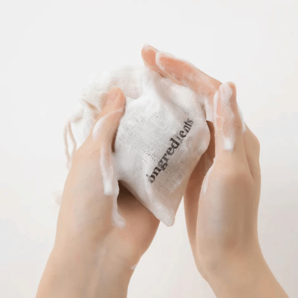 Ręce pieniące Óngredients Jeju Green Tea Cleansing Ball w materiałowej torebce – wegańskie oczyszczanie twarzy z zieloną herbatą.