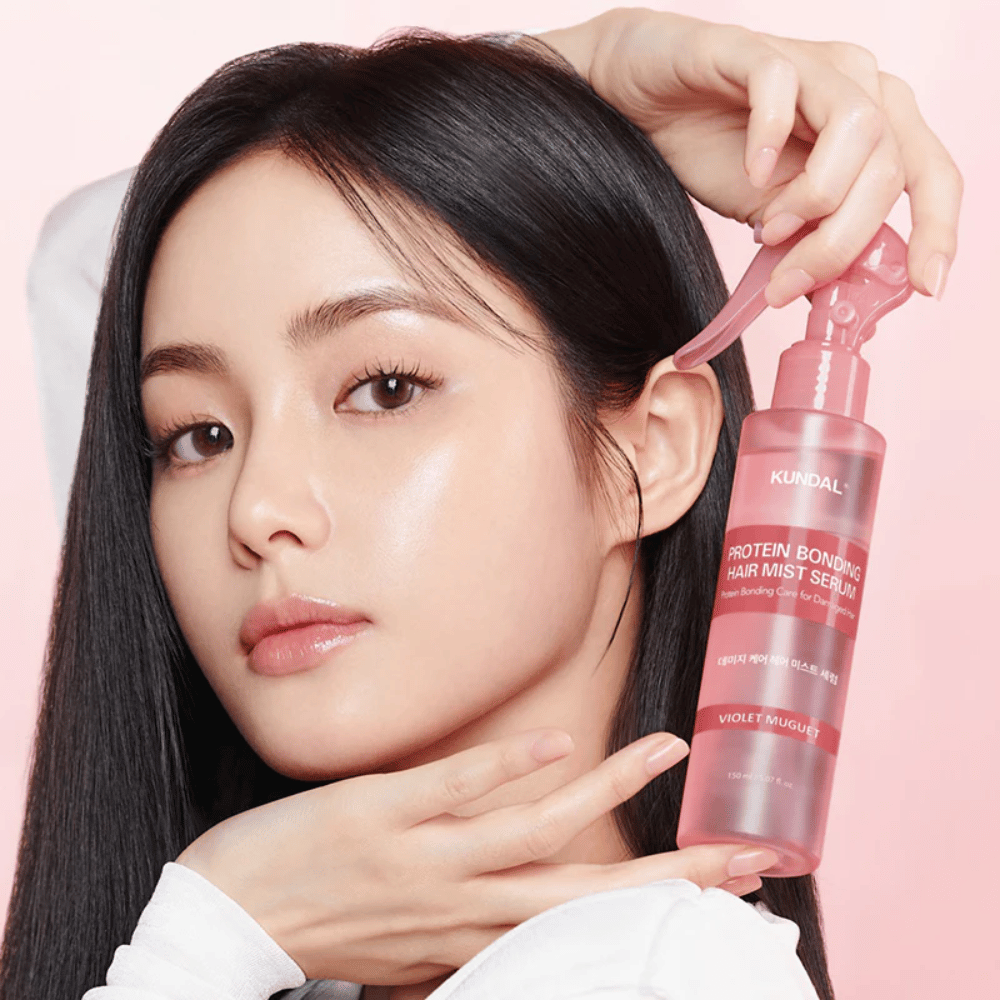 Kobieta z długimi czarnymi włosami trzyma KUNDAL Protein Bonding Hair Mist Serum Violet Muguet przed twarzą, na tle jasnoróżowym.