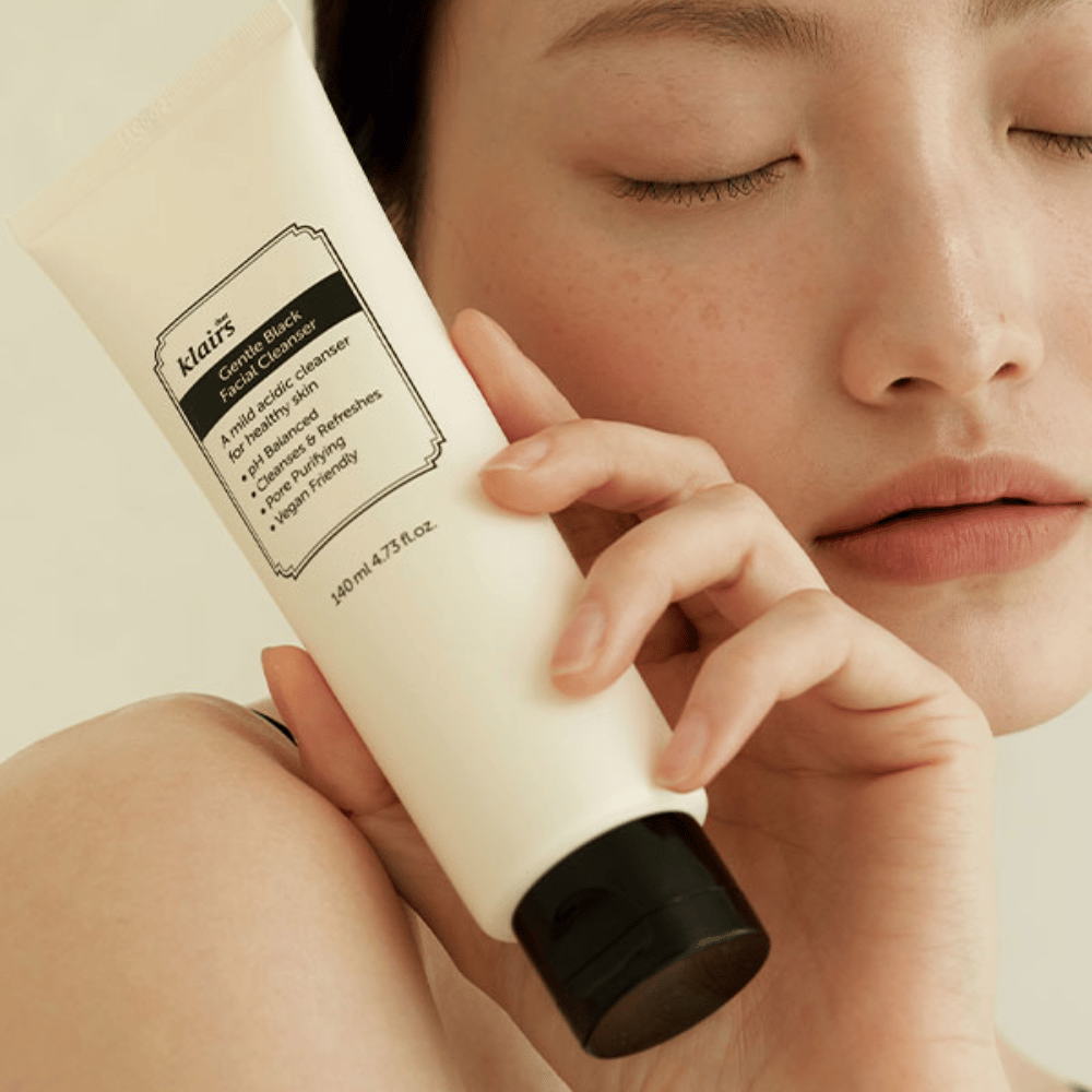 Osoba trzyma tubę Klairs Gentle Black Facial Cleanser przy swoim policzku, zamknięte oczy i naturalny odcień skóry widoczne w tle.