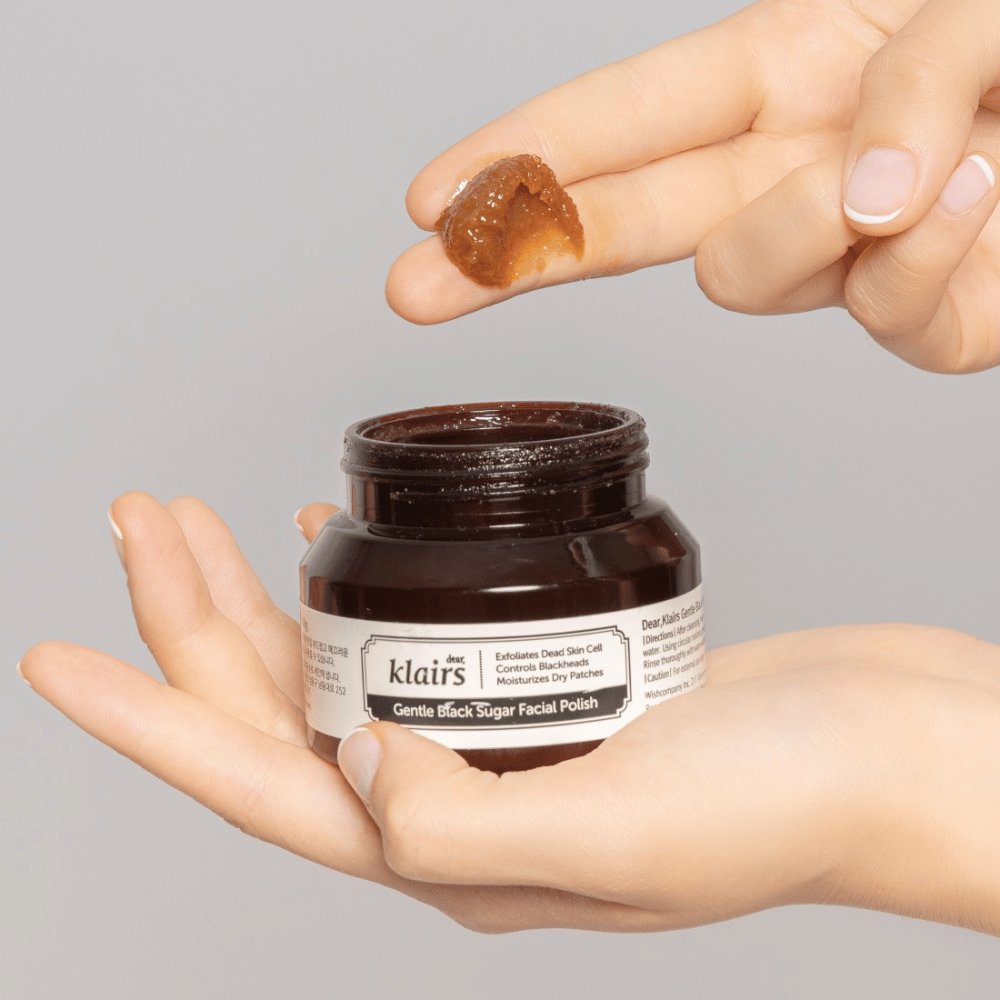 Ręce trzymają otwarty słoik Klairs Gentle Black Sugar Facial Polish i pokazują produkt na palcu, na szarym tle.
