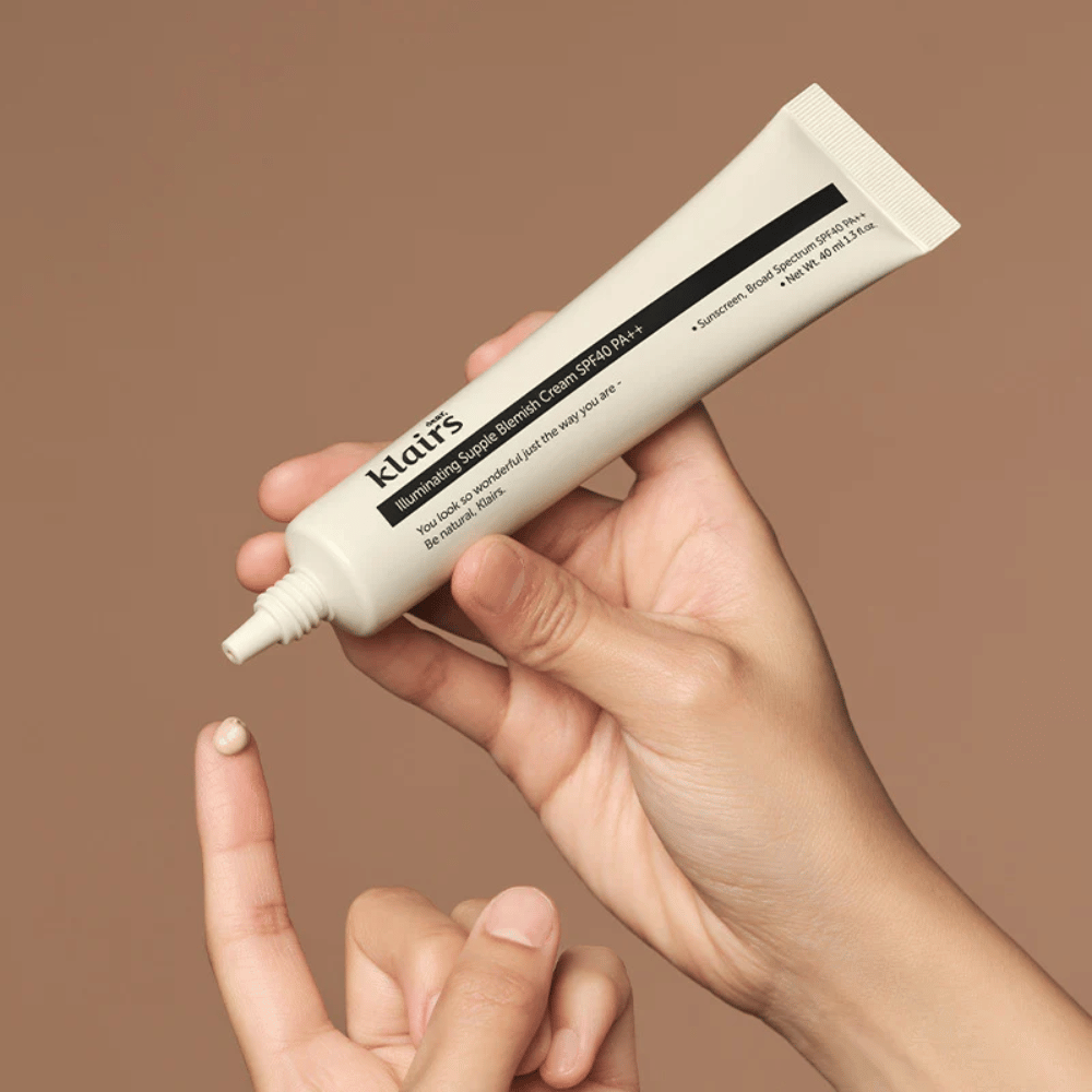 Ręka trzyma tubę Klairs Illuminating Supple Blemish Cream SPF40 PA++ i wyciska niewielką ilość kremu na palec, na brązowym tle.