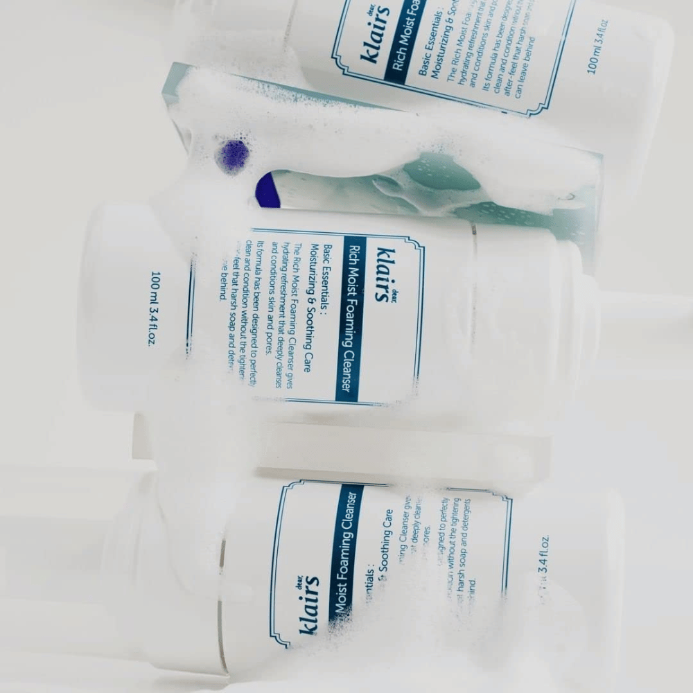 Trzy butelki Klairs Rich Moist Foaming Cleanser pokryte pianką i kroplami wody, umieszczone na białej powierzchni.