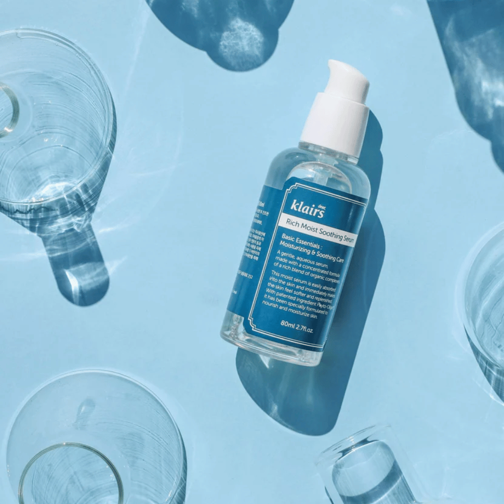 Butelka Klairs Rich Moist Soothing Serum wśród szklanek z wodą z refleksami na niebieskim tle.