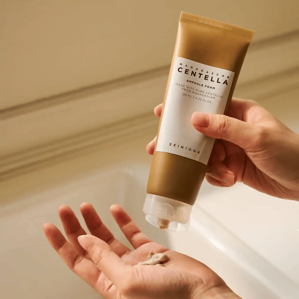 Produkt na obrazku to "Madagascar Centella Ampoule Foam" od SKIN1004. Jest to pianka do mycia twarzy, która została stworzona z Centelli z Madagaskaru. Zawiera 125 ml / 4.22 fl.oz. Ręka wyciska niewielką ilość produktu do użycia.
