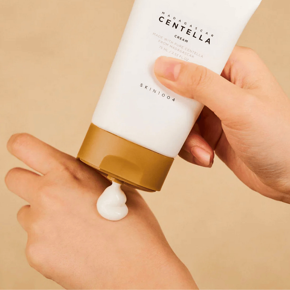 Obrazek przedstawia osobę trzymającą tubkę "Madagascar Centella Cream" od SKIN1004. Jest to krem do skóry wyprodukowany z czystej Centelli z Madagaskaru, o pojemności 75 ml / 2.53 fl.oz. Krem jest nakładany na rękę i ma na celu dostarczenie składników odżywczych i nawilżenie skóry.