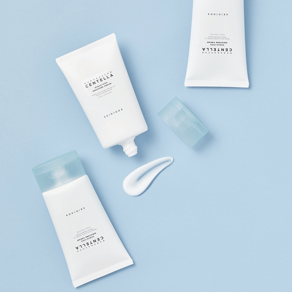 Dwie białe tuby z SKIN1004 produktami do pielęgnacji skóry Centella, odrobina produktu i przezroczysta niebieska nakrętka, na jasnoniebieskim tle.