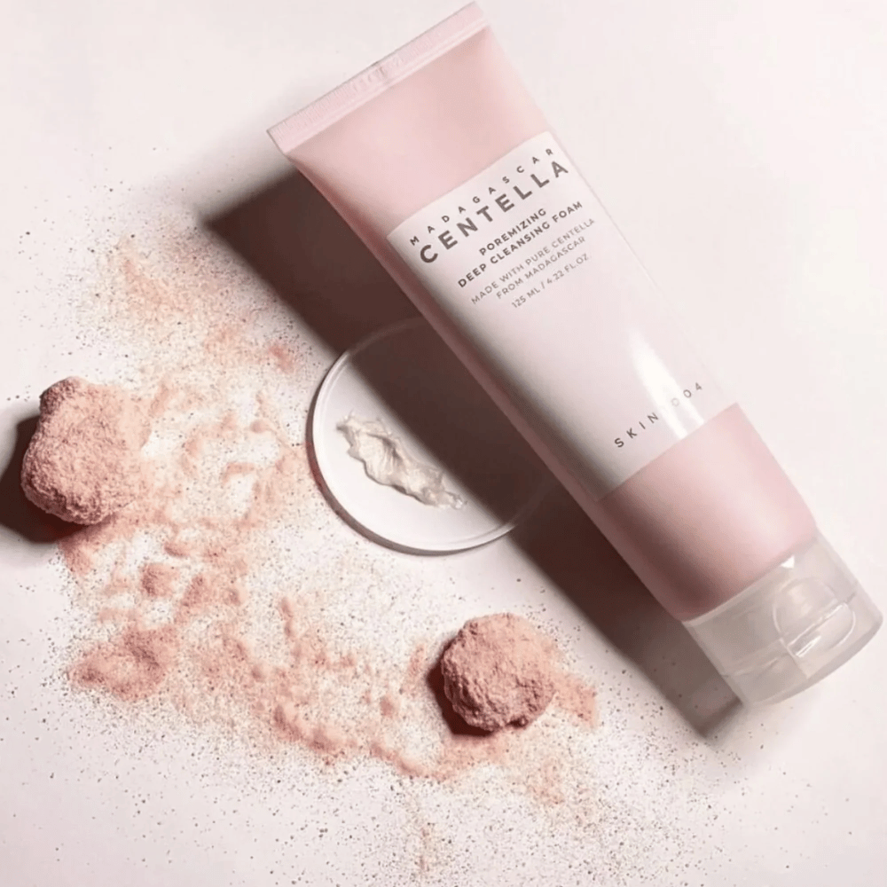 Madagascar Centella Poremizing Deep Cleansing Foam jest prezentowana w różowej tubie. Tuba leży na jasnym podłożu wraz z różowym proszkiem, który daje poczucie czystości i świeżości. Produkt jest zaprojektowany do głębokiego oczyszczania skóry za pomocą czystej centelli z Madagaskaru.