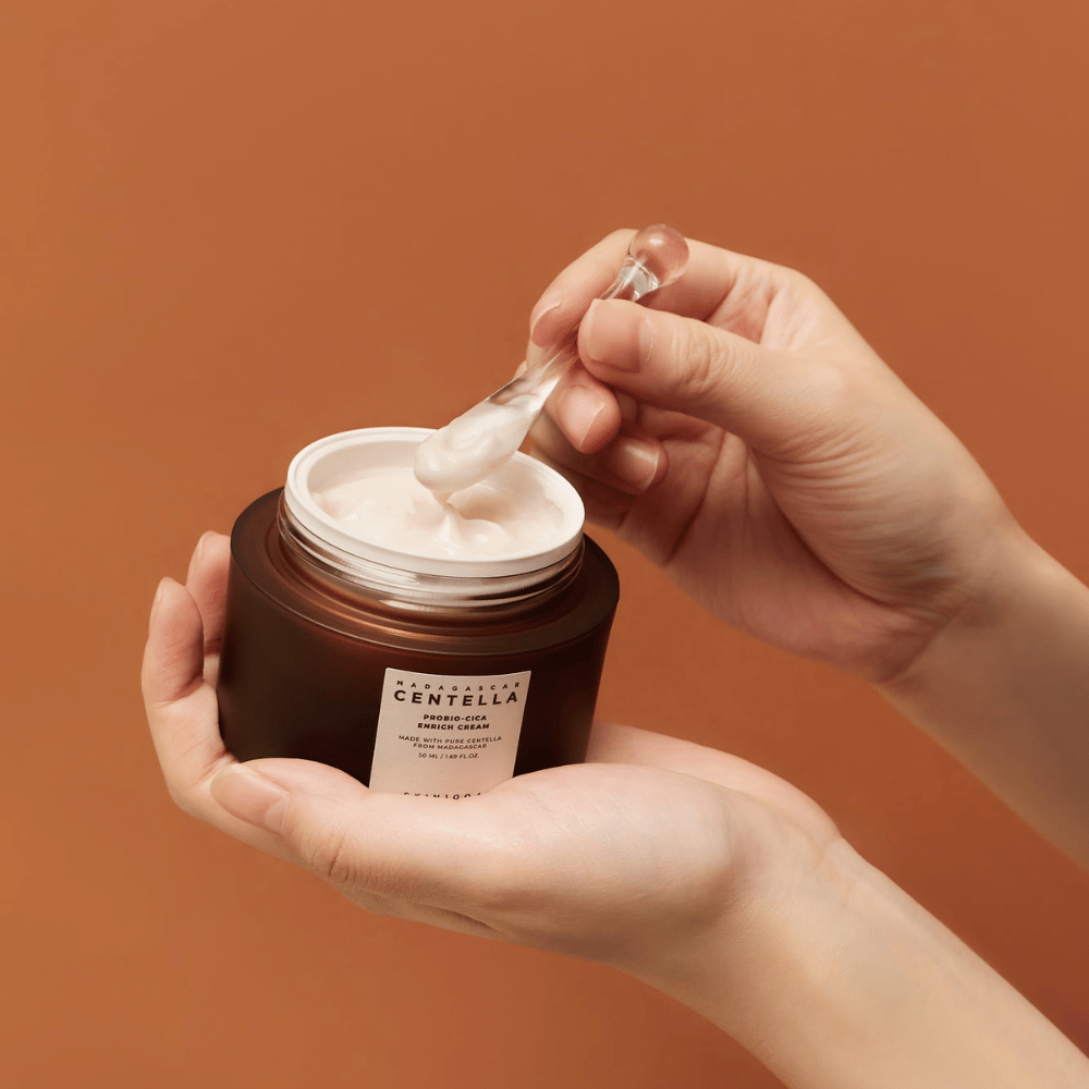 Ręce trzymające otwarty słoik Madagascar Centella Probio-Cica Enrich Cream i szpatułkę z kremem na jednolitym terakotowym tle.