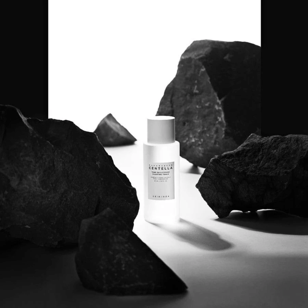 Biała butelka SKIN1004 Madagascar Centella Tone Brightening Boosting Toner  umieszczona dramatycznie wśród dużych czarnych kamieni na białym tle.