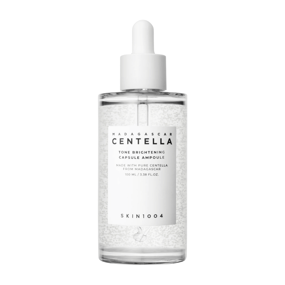 Obrazek przedstawia butelkę "SKIN1004 Madagascar Centella Tone Brightening Capsule Ampoule". Jest to przezroczysta butelka z białym aplikatorem kroplowym. Na etykiecie jest napisane, że produkt zawiera czyste składniki centella z Madagaskaru. Zawartość butelki wygląda na to, że ma małe kapsułki lub cząsteczki, co może być częścią jej rozjaśniających właściwości pielęgnacyjnych. Butelka zawiera 100 ml / 3.38 fl.oz. produktu.