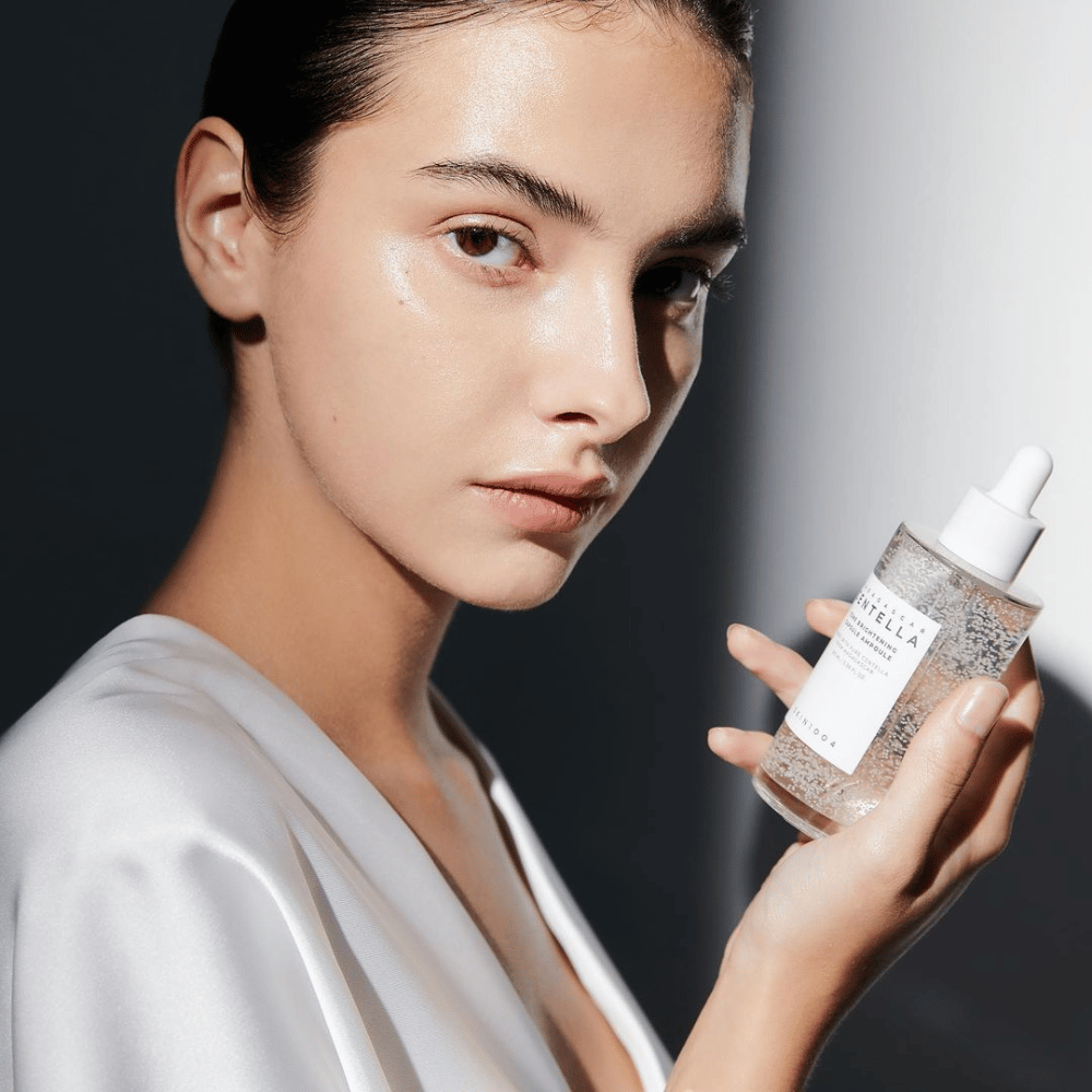 Na obrazku widać osobę trzymającą butelkę z aplikatorem kroplowym. Butelka jest oznaczona "SKIN1004 Madagascar Centella Tone Brightening Capsule Ampoule". Osoba ma lśniącą tonację skóry, która odbija światło, co sugeruje możliwy efekt działania produktu.