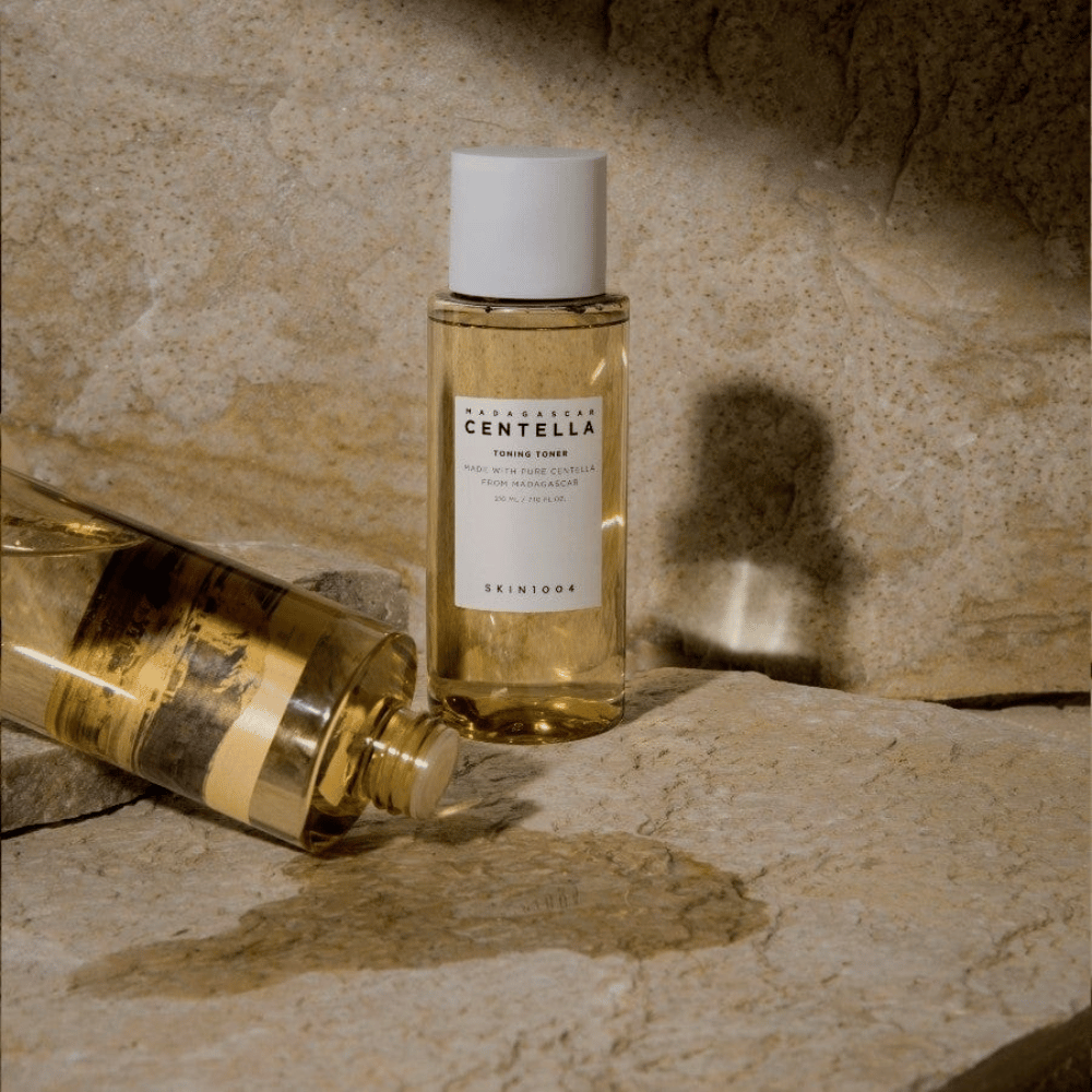Obraz przedstawia butelkę "Madagascar Centella Toning Toner" od SKIN1004. Jest to przezroczysta butelka wypełniona żółtawym płynem, prawdopodobnie tonikiem do pielęgnacji skóry. Napisano, że jest wykonana z czystej centelli z Madagaskaru, a zawartość to 210 ml / 7.10 fl.oz. Butelka i leżąca butelka są umieszczone na kamienistej powierzchni, co daje naturalne i minimalistyczne odczucie.