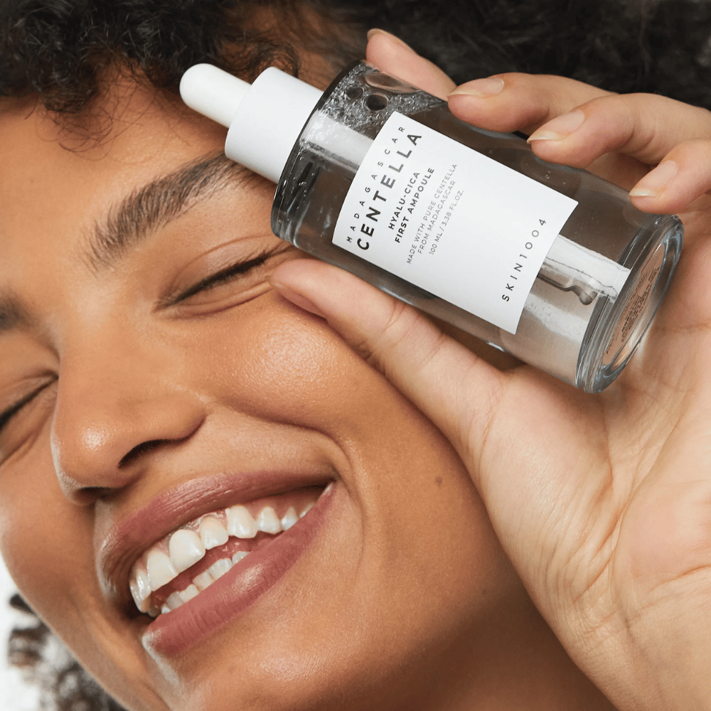 Uśmiechnięta kobieta trzyma Skin1004 Madagascar Centella Hyalu-Cica First Ampoule obok twarzy.