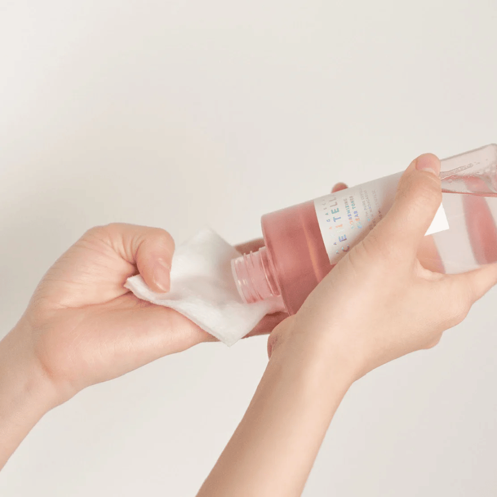 Osoba wylewa Madagascar Centella Poremizing Clear Toner z przezroczystej butelki na wacik. Produkt, który ma różowy odcień, jest używany do odświeżania i oczyszczania skóry. Widoczne są ręce, co pokazuje aplikację toniku.